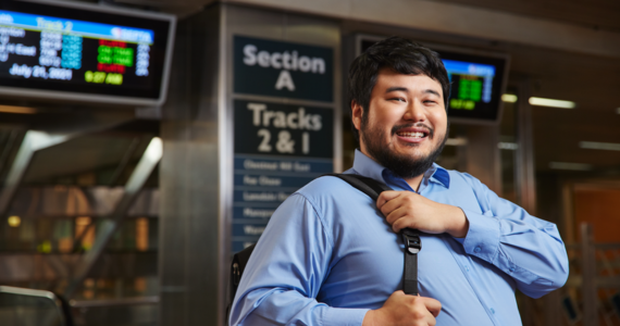 Man with dark hair and blue collared shirt holding work bag waiting by train platform